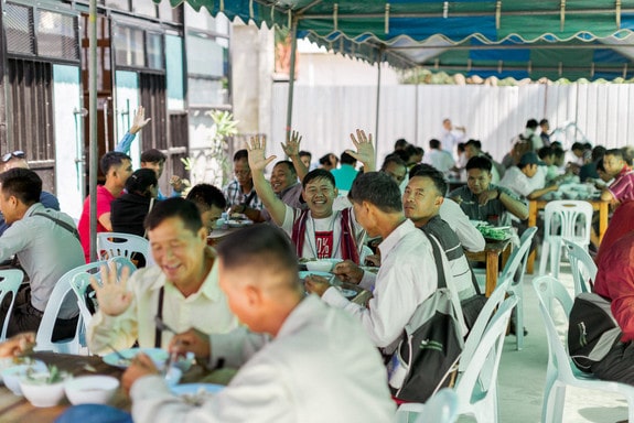 People sitting around tables, eating meal and waving to the camera