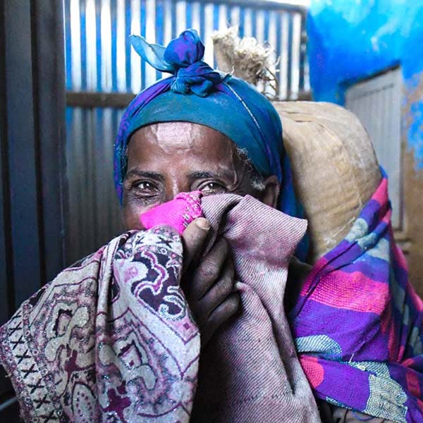An aged African woman covering her face with clothes, and carrying some belongings on back