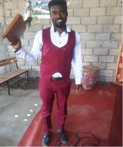 Pastor of Dawn's sponsored church in East Africa appearing in the photo with a red suit vest, suit pants, with a white button up shirt holding a Bible and standing at the church.