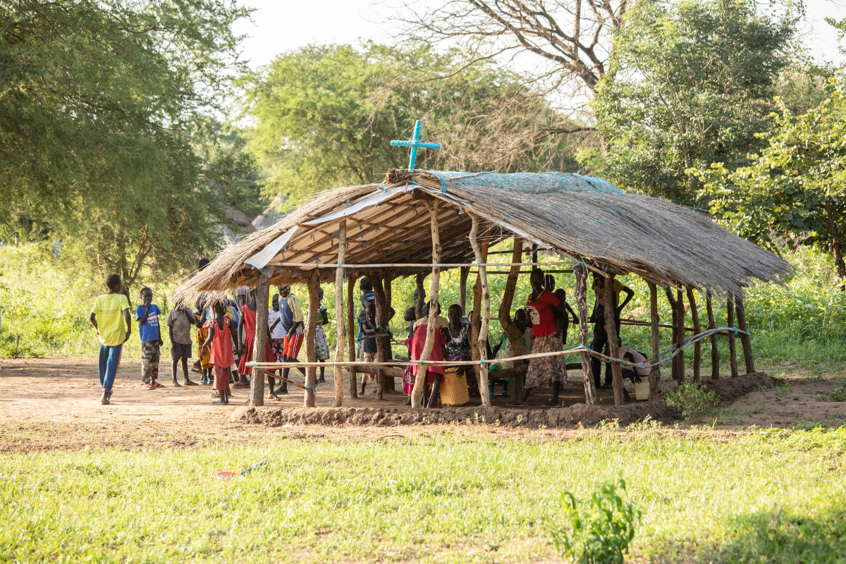 Church plant in South Sudan, structure made up of sticks with a blue cross atop in and a group of about 50 people gathered below.