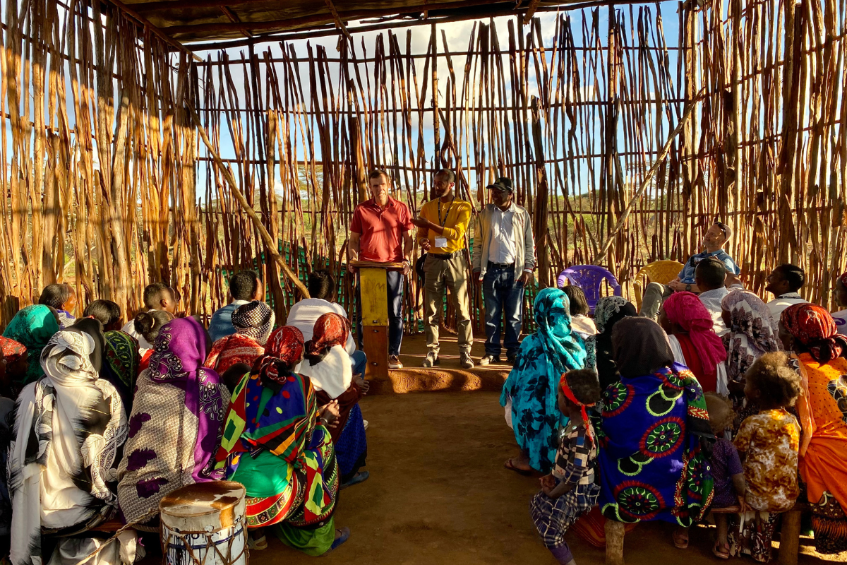 Ray Noah speaking to a church in a rural village in Ethiopia, structure is made up of sticks, people are primarily women and girls in cultural dress.